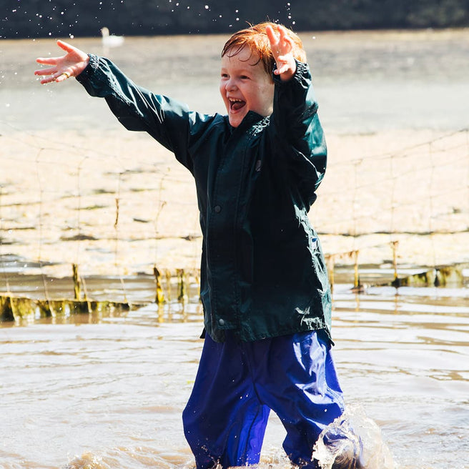 A happy ginger boy in the Originals Waterproof Jacket Green, paddling in water with arms raised and a big grin.